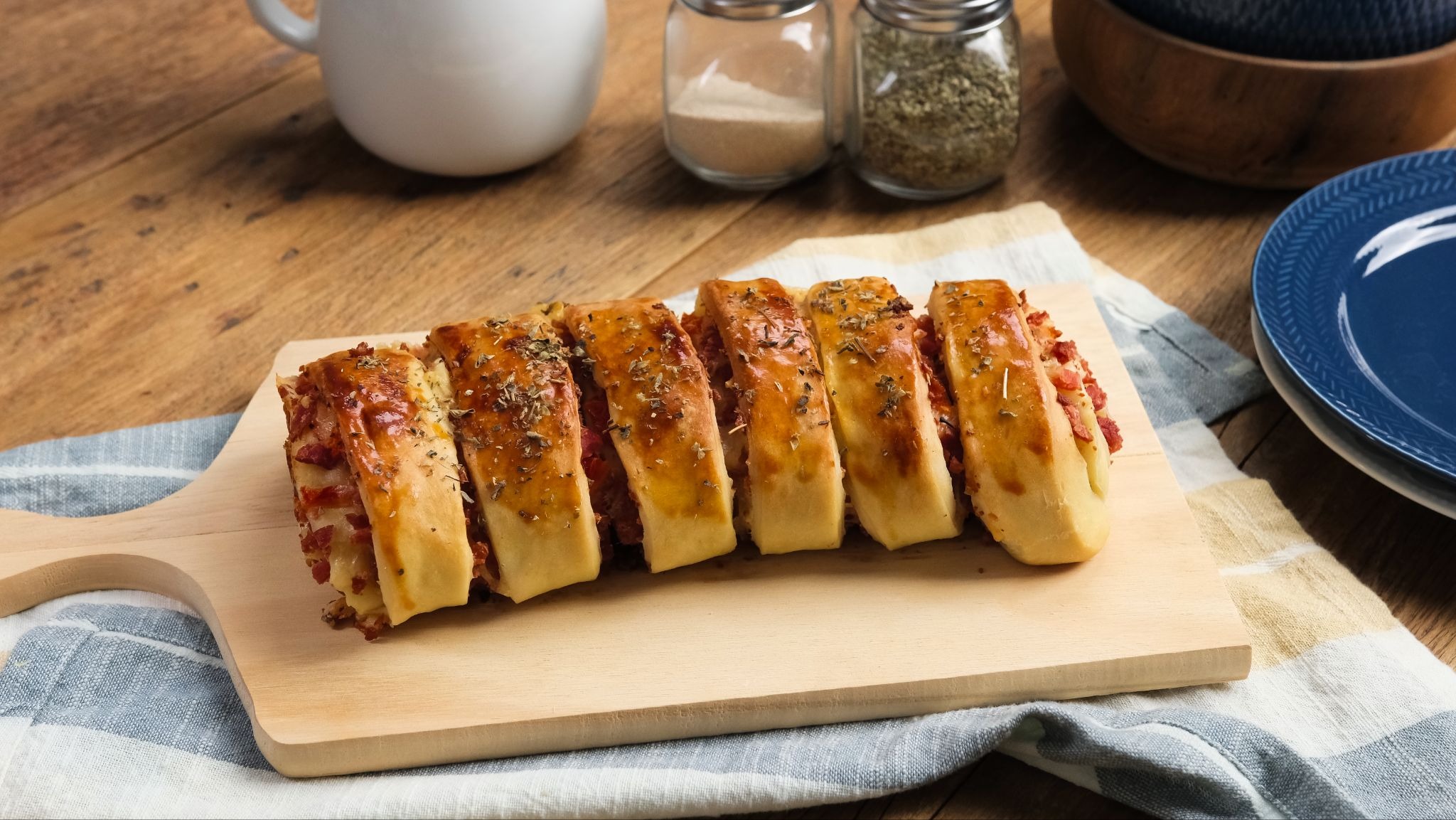 O lanche perfeito: receita de pão caseiro recheado. Pão recheado com bacon e queijo, cortado em fatias e polvilhado com orégano, sobre uma tábua de madeira clara. Ao fundo, uma caneca branca, potes de temperos e um prato azul em uma mesa de madeira
