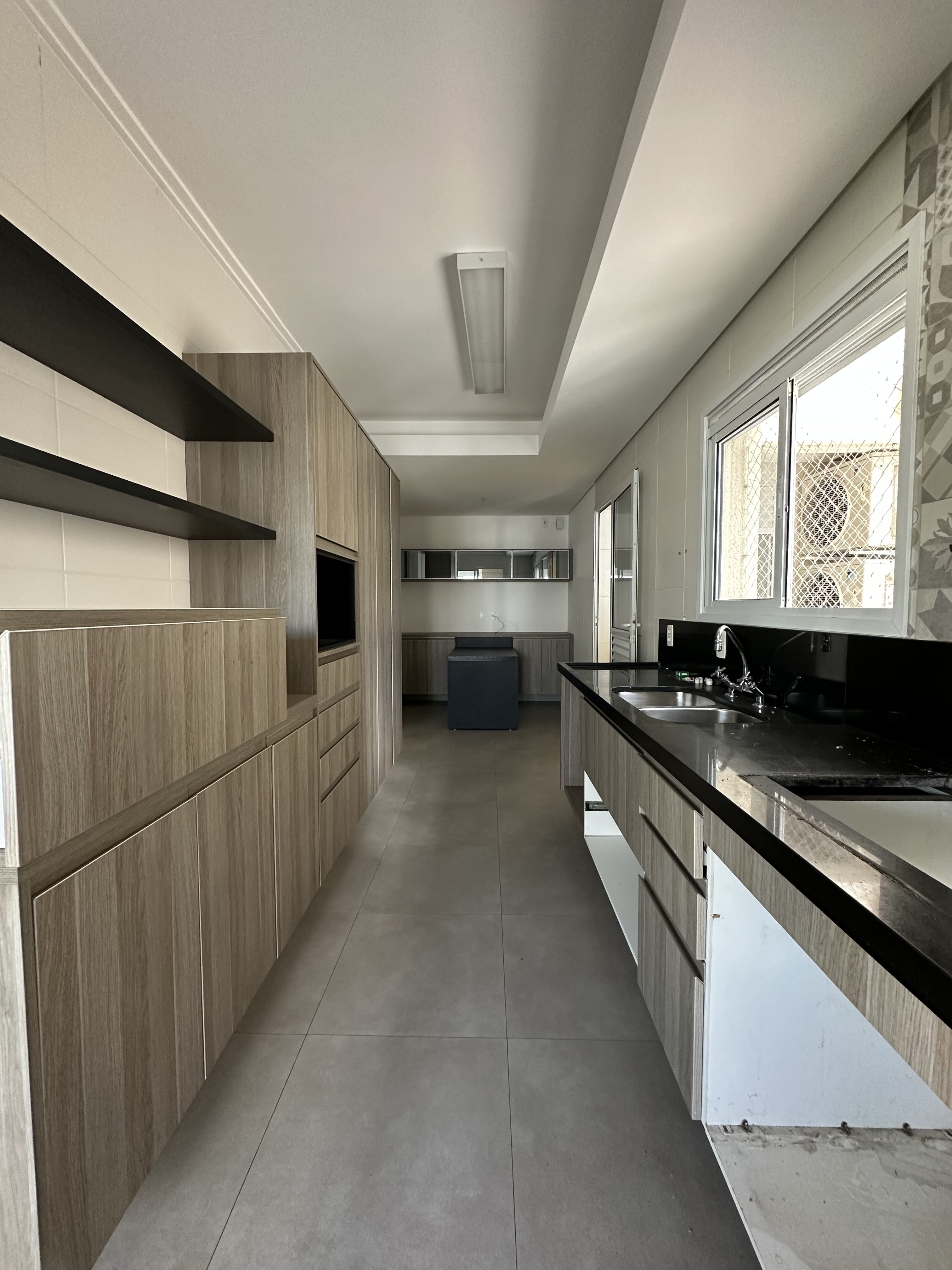 Cozinha comprida e estreita com armários de madeira clara e bancadas pretas, pia dupla de inox, prateleiras escuras na parede esquerda e uma janela com grade à direita. Ao fundo, uma máquina de lavar escura e armários suspensos. O piso é de azulejos cinza claro e o teto tem iluminação retangular.