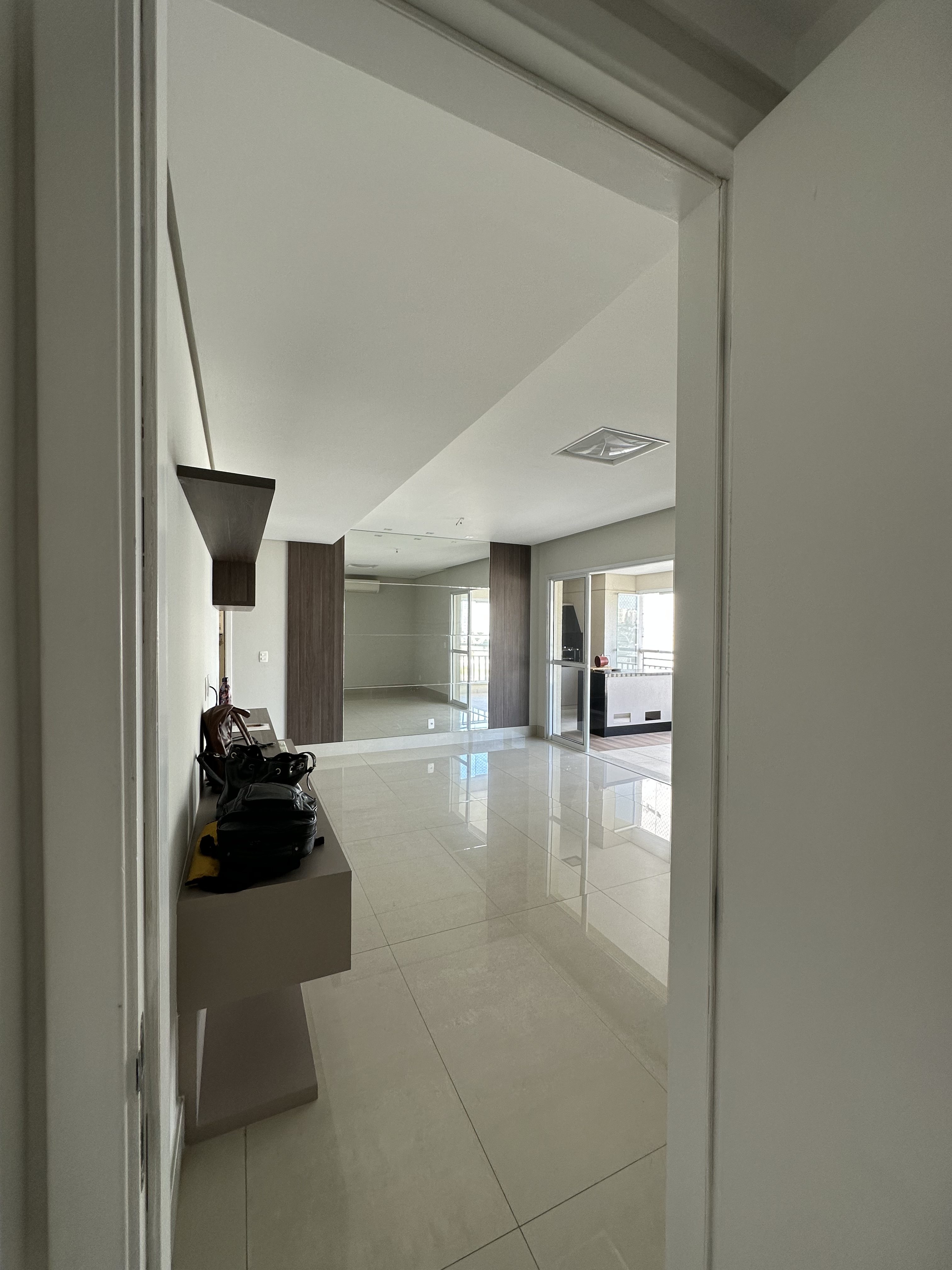 Vista de um corredor estreito para uma sala de estar vazia com piso de porcelanato branco brilhante. À esquerda, um aparador bege com uma bolsa preta e um objeto marrom. A parede do fundo tem um espelho grande que reflete a sala e uma porta de vidro que leva a uma varanda. O teto é branco com uma luminária quadrada. A iluminação é clara e natural