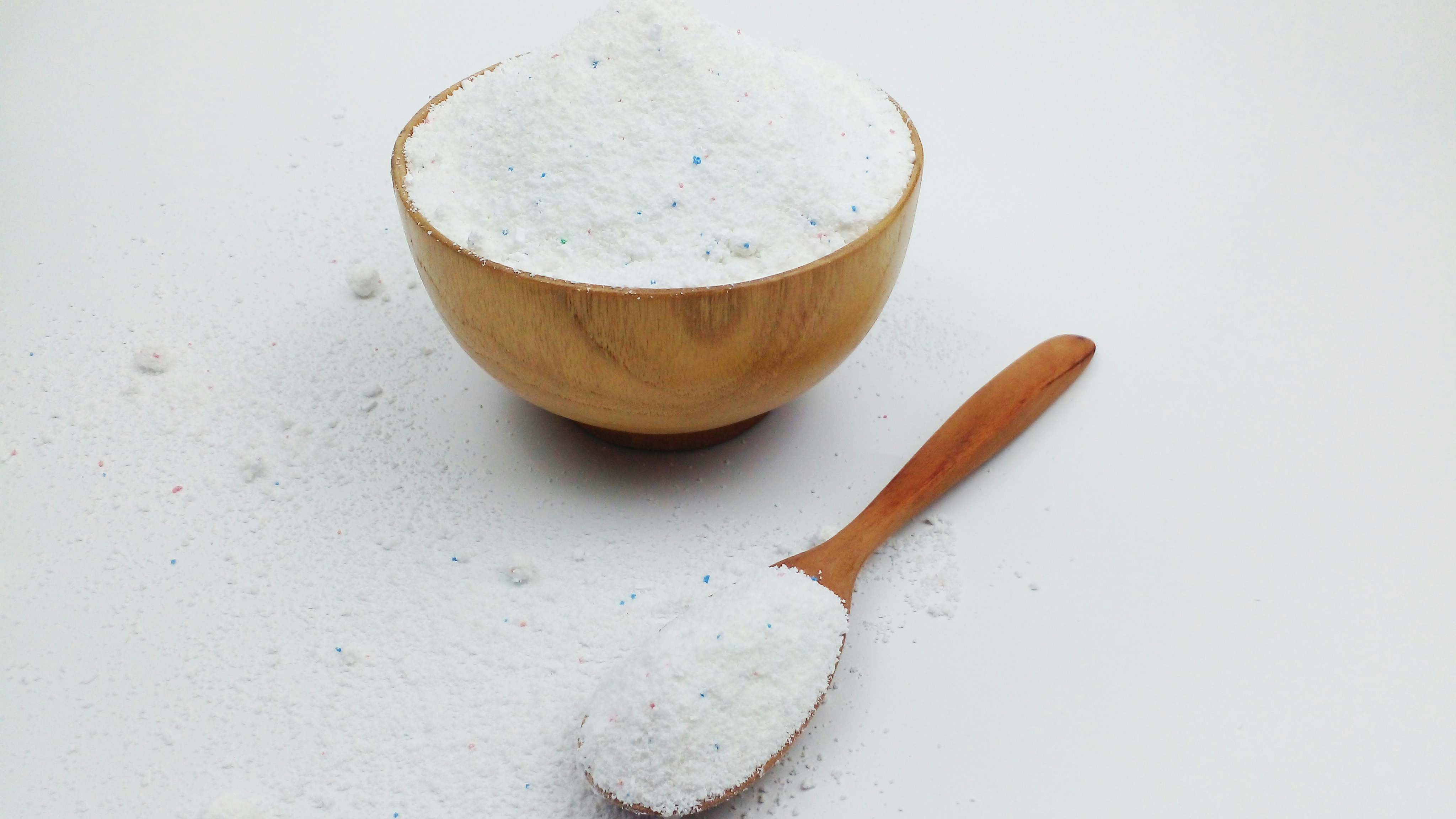 Pó branco com grânulos azuis e vermelhos em uma tigela de madeira e em uma colher de pau, sobre uma superfície branca com respingos