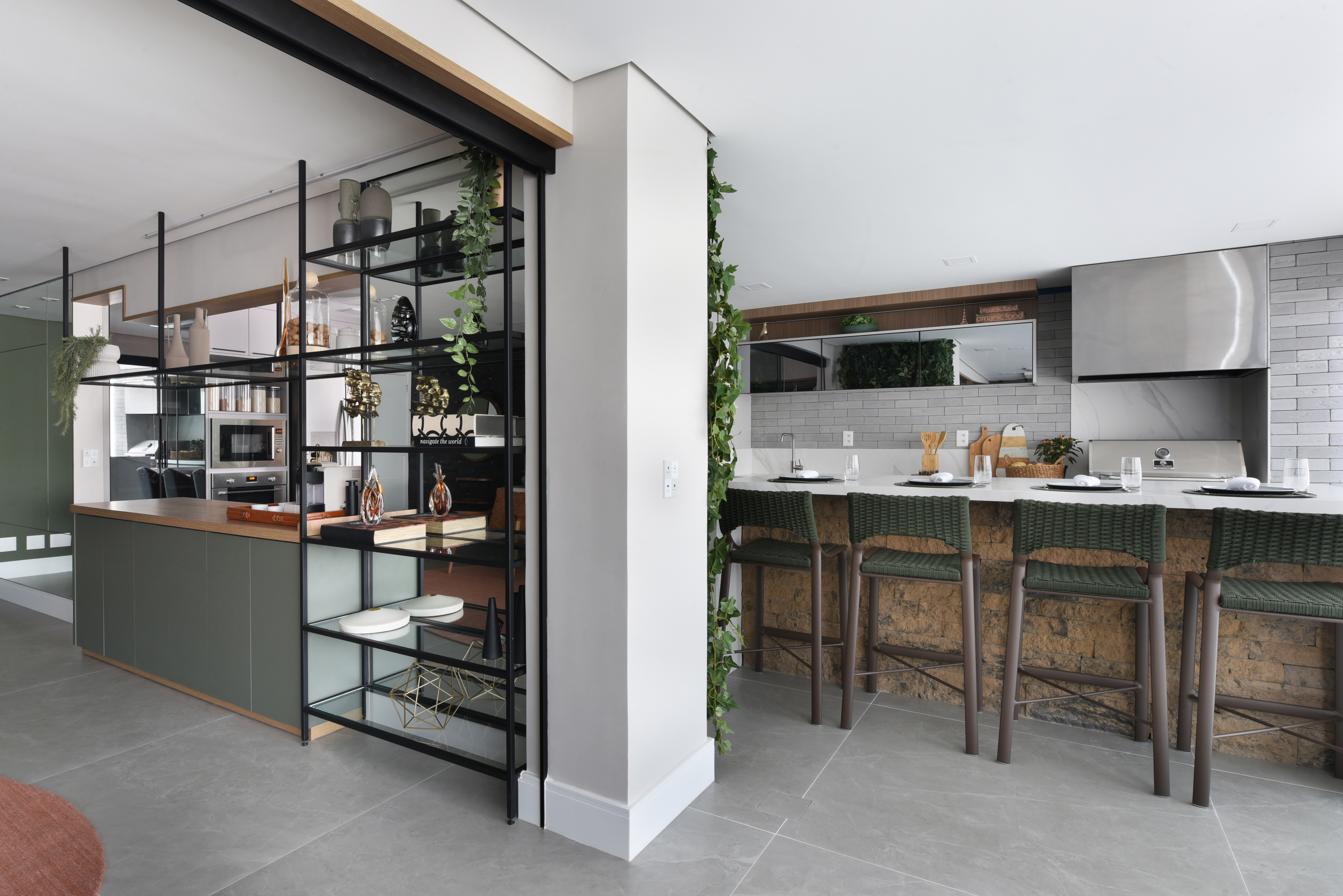 Com mais formatos e funções, estantes mantêm protagonismo nos interiores. Projeto de Rosângela Pena. Cozinha moderna com ilha central e bancada de refeições. À esquerda, armários verdes e prateleiras pretas com objetos decorativos e plantas pendentes. À direita, bancada branca com quatro banquetas de vime verde, parede de tijolos rústicos e eletrodomésticos de aço inoxidável.