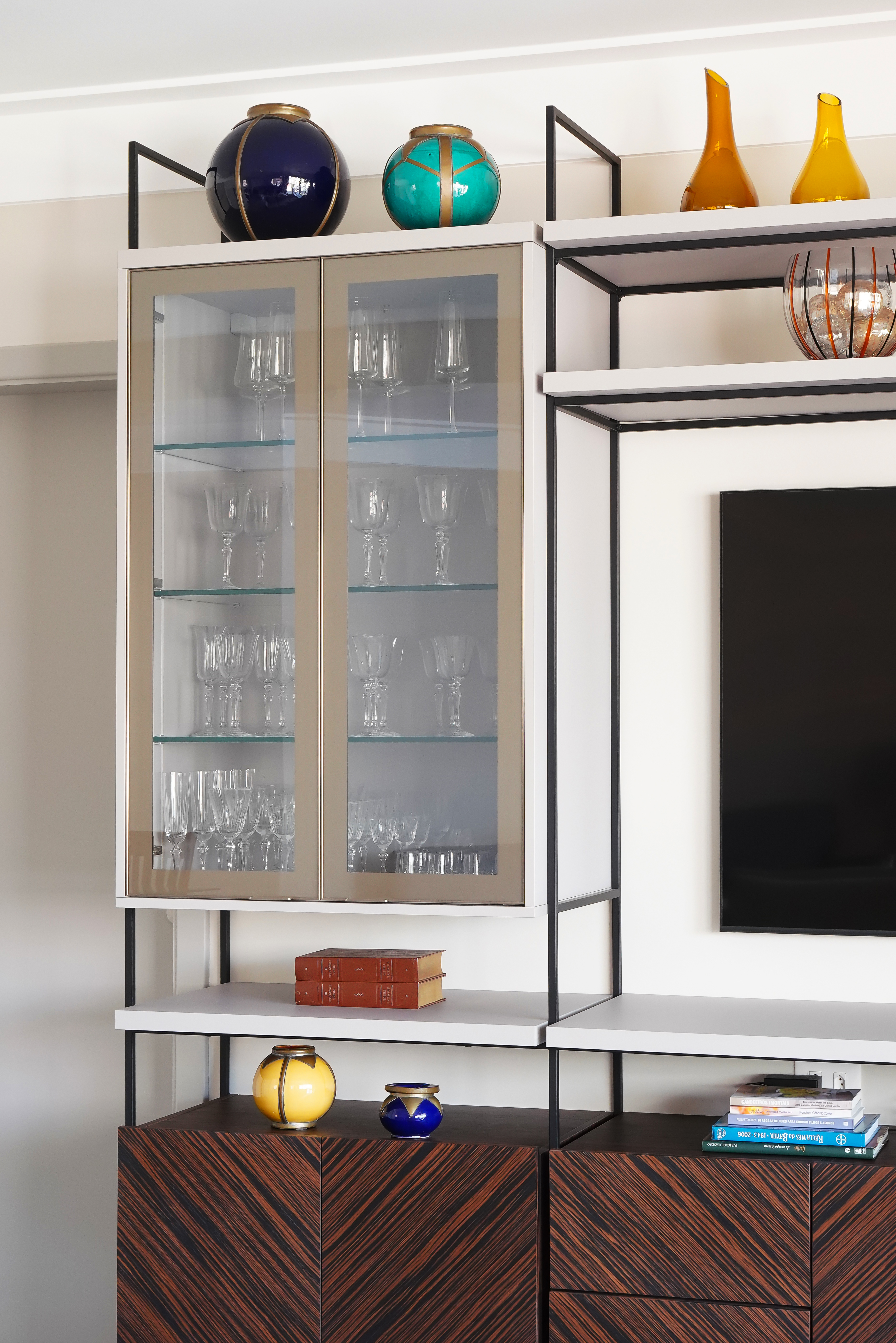 Com mais formatos e funções, estantes mantêm protagonismo nos interiores. Projeto de Rosângela Pena. Estante modular moderna com armário superior de vidro exibindo taças, prateleiras com livros e vasos decorativos coloridos, e base de madeira escura com padrão diagonal