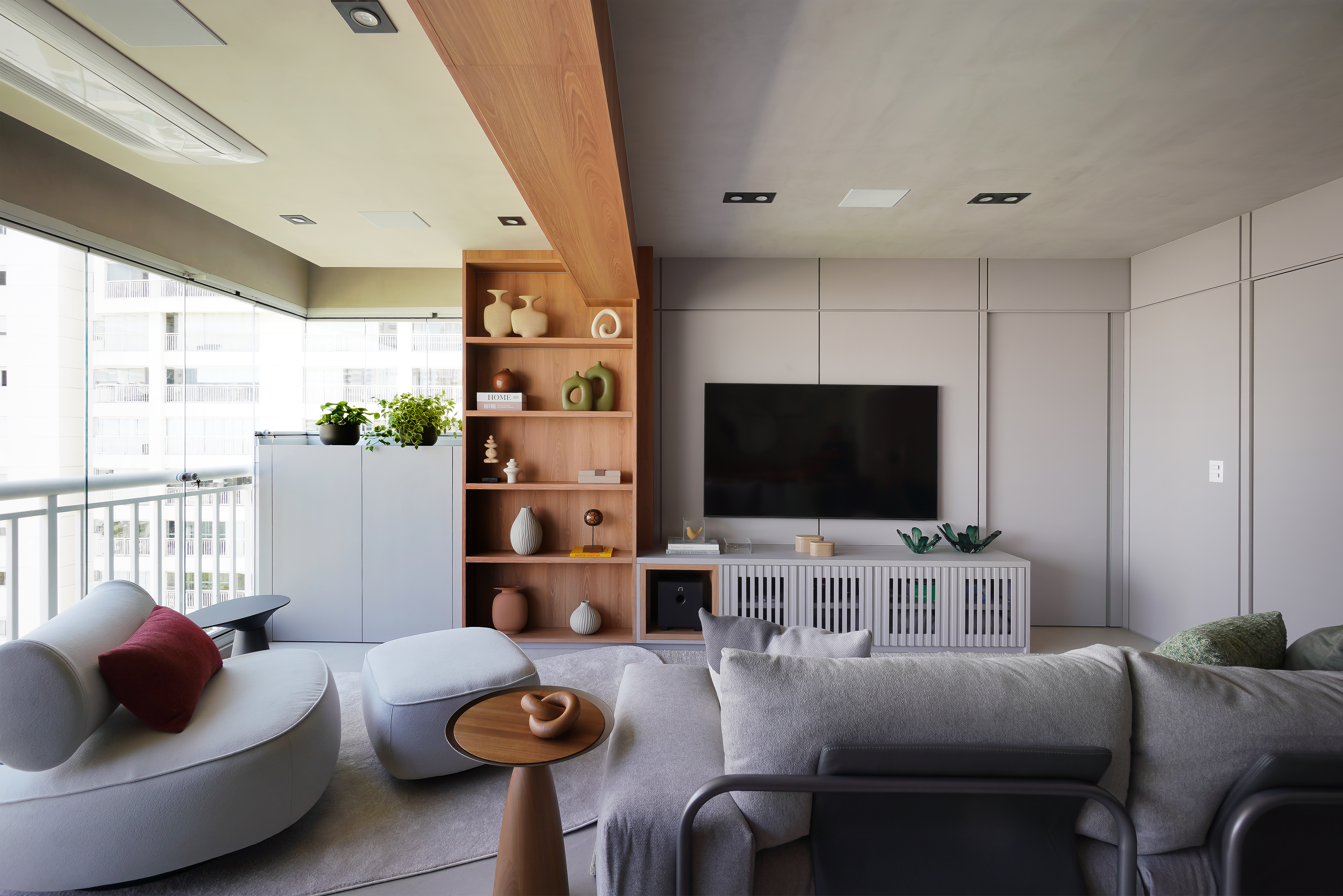 Com mais formatos e funções, estantes mantêm protagonismo nos interiores. Projeto de Rosângela Pena. Sala de estar moderna com sofá cinza, poltrona branca com almofada vermelha, estante de madeira clara com objetos decorativos e televisão embutida. Há um balcão branco com plantas e uma varanda envidraçada ao fundo, revelando prédios.